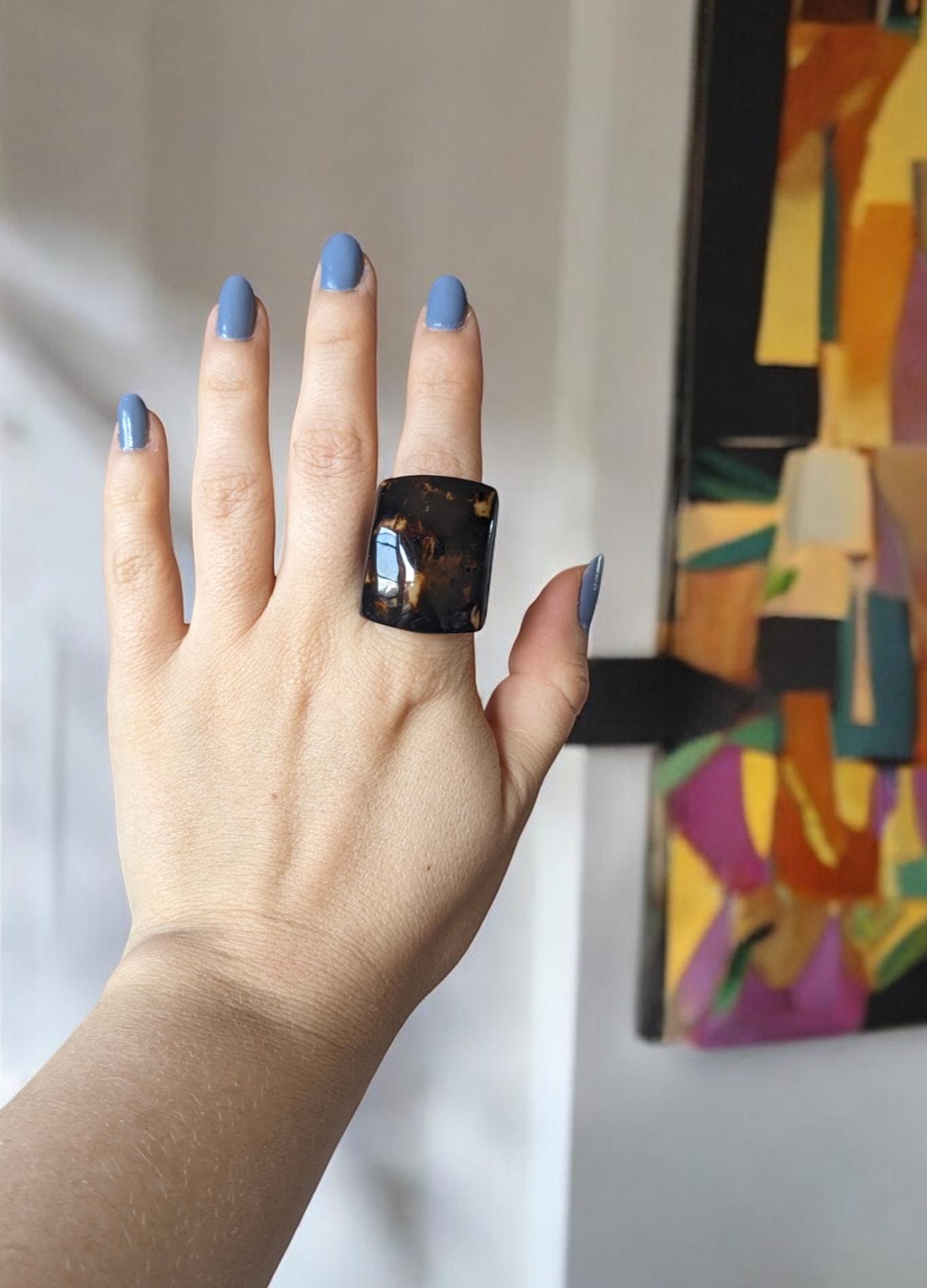 Hand with a large black ring and blue nail polish in front of a colorful abstract painting.