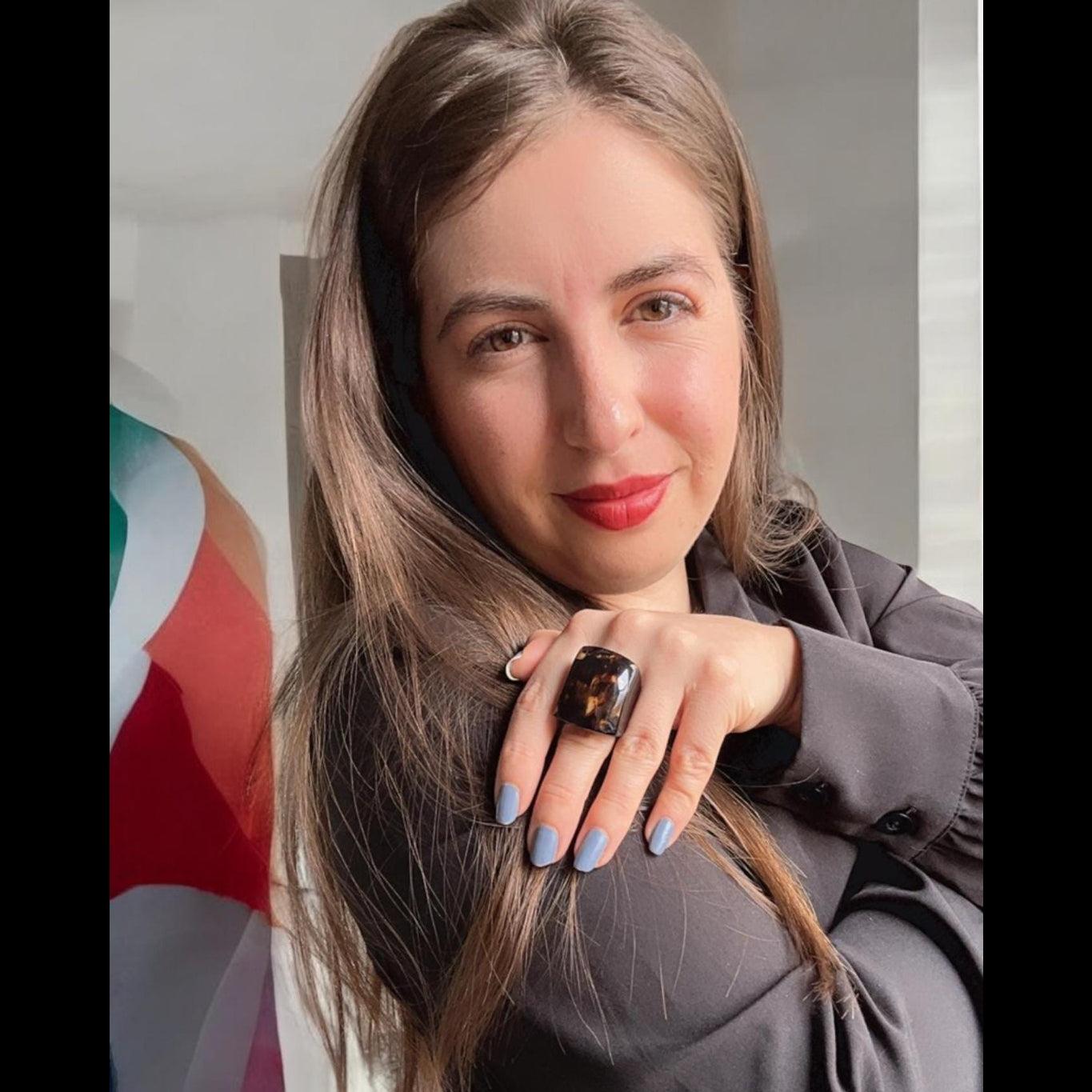 Woman with long hair and a ring, standing indoors with a colorful abstract painting in the background.