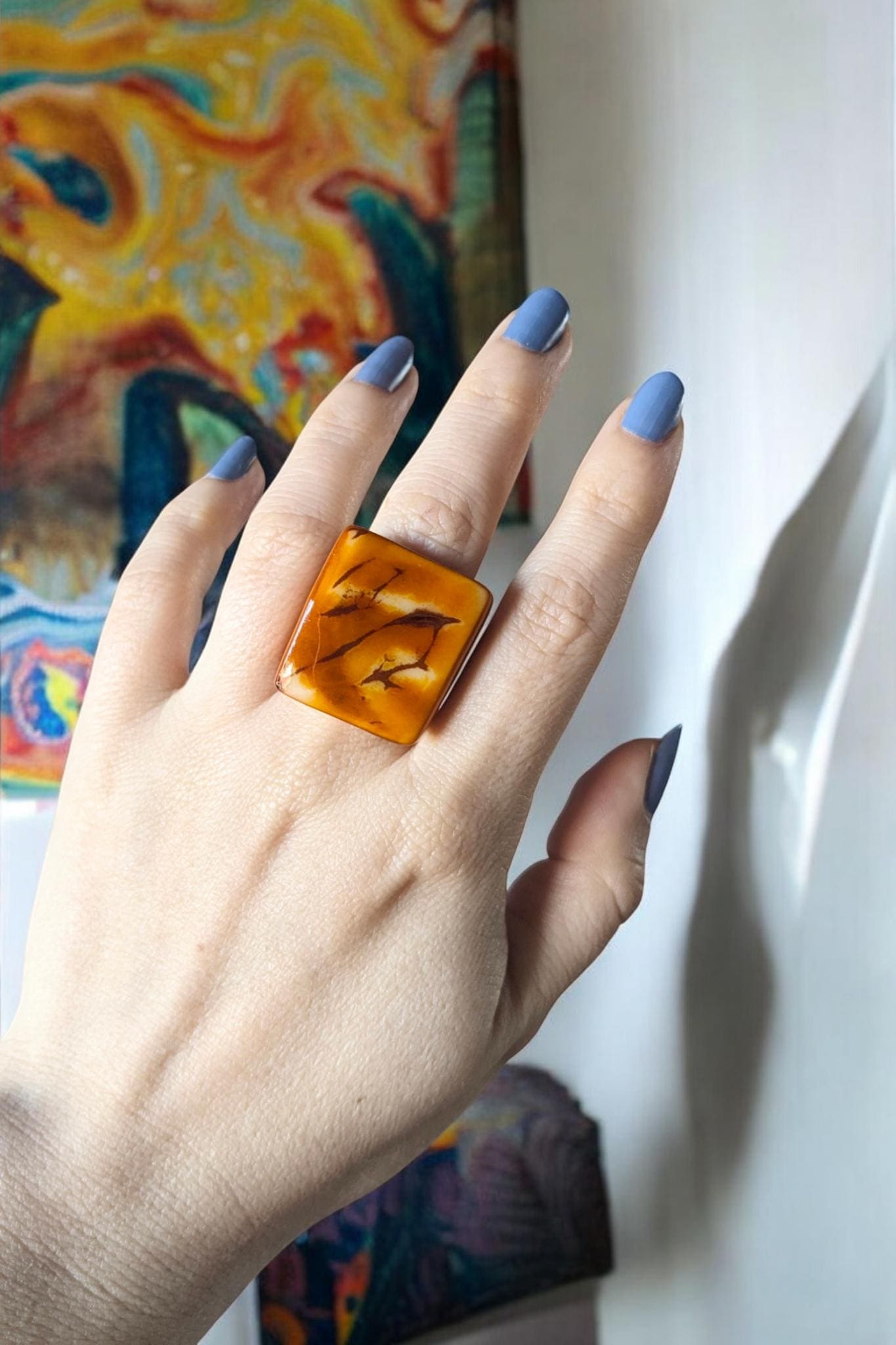 Hand wearing a large canary yellow ring  with blue nail polish against a colorful abstract background